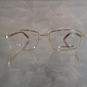 May include: Gold-rimmed spectacles with a square frame. The brand name "VERSACE" is printed on the right lens.
