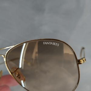 Puede incluir: Gafas de sol aviador doradas con lentes tintados de color marr&oacute;n. Las gafas tienen la palabra "FANTASEES" impresa en la lente derecha.
