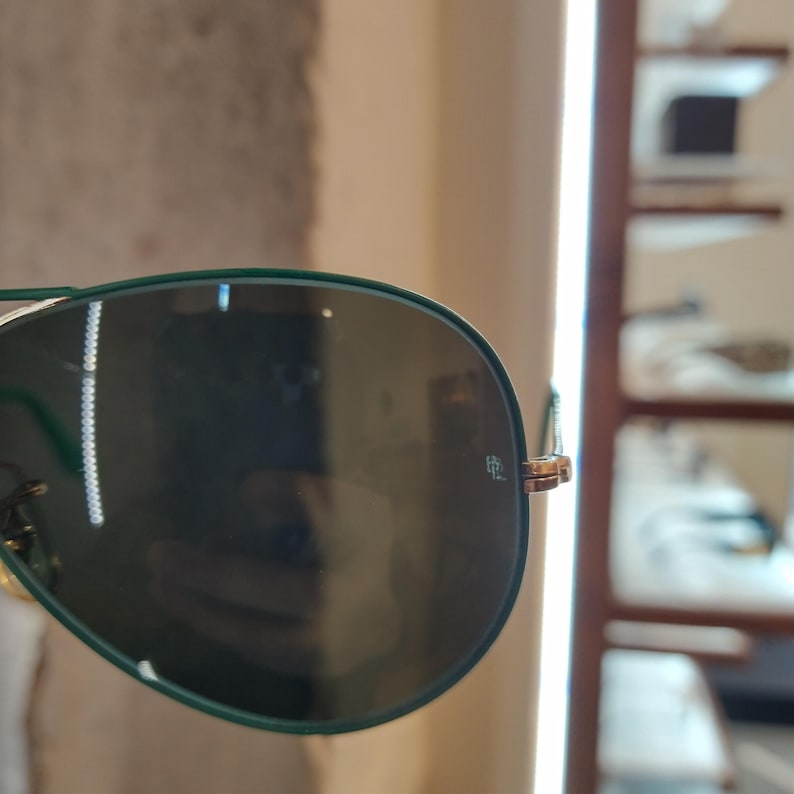 Op de afbeelding: Close-up van een groene aviator zonnebril met een gouden montuur. De zonnebril heeft een reflecterende lens en het Ray-Ban logo is zichtbaar op de rechter lens.