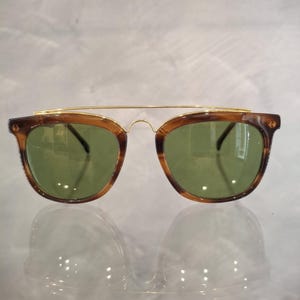May include: Sunglasses with a brown tortoiseshell frame and green lenses. The sunglasses have a gold-colored metal bridge connecting the lenses. The lenses are square-shaped with rounded edges.