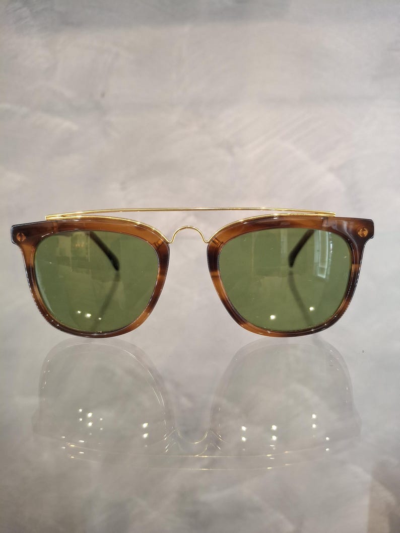 May include: Sunglasses with a brown tortoiseshell frame and green lenses. The sunglasses have a gold-colored metal bridge connecting the lenses. The lenses are square-shaped with rounded edges.
