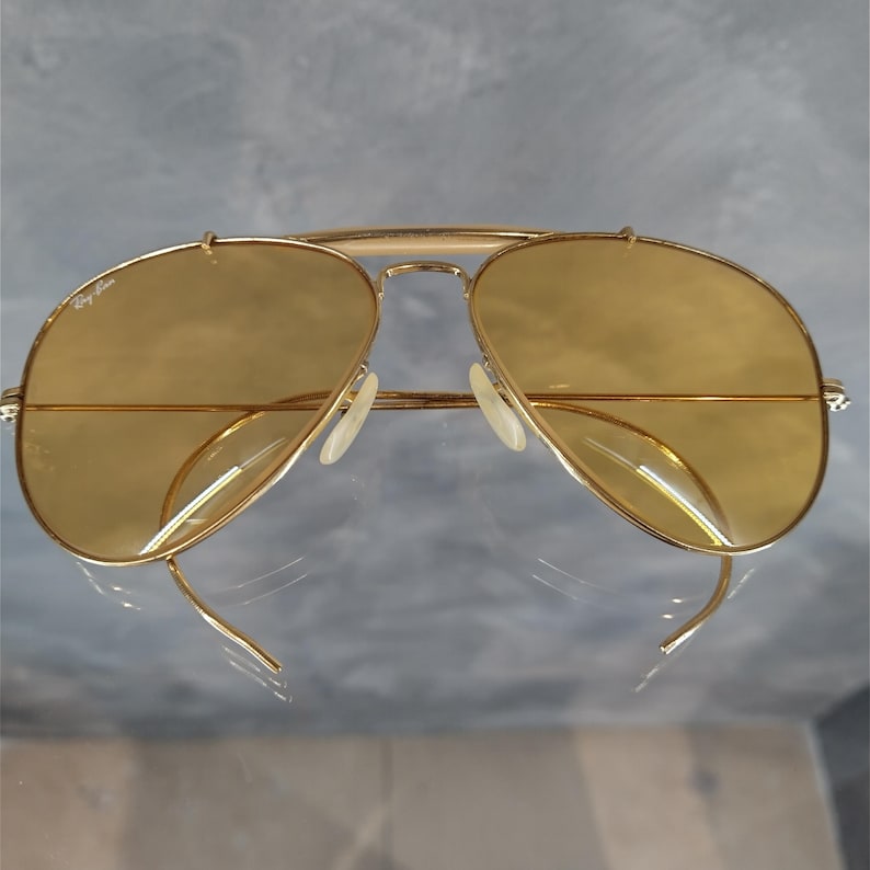 May include: Gold-framed aviator sunglasses with amber-tinted lenses. The sunglasses feature a double bridge and a classic teardrop shape. The Ray-Ban logo is visible on the upper left lens. The sunglasses are displayed on a reflective surface.