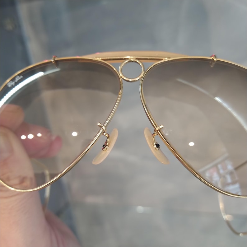 May include: Gold aviator sunglasses with brown tinted lenses. The sunglasses have a metal frame and a nose bridge with a circular detail.