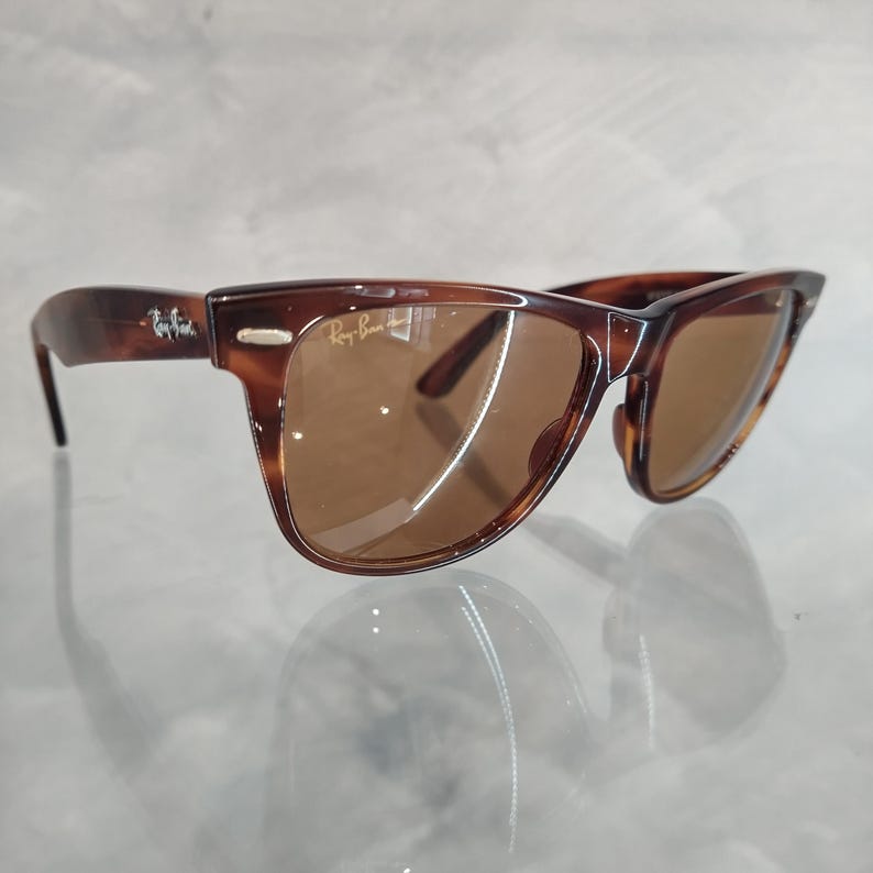 May include: Brown tortoise shell Ray-Ban Wayfarer sunglasses with brown lenses. The sunglasses are on a white surface.
