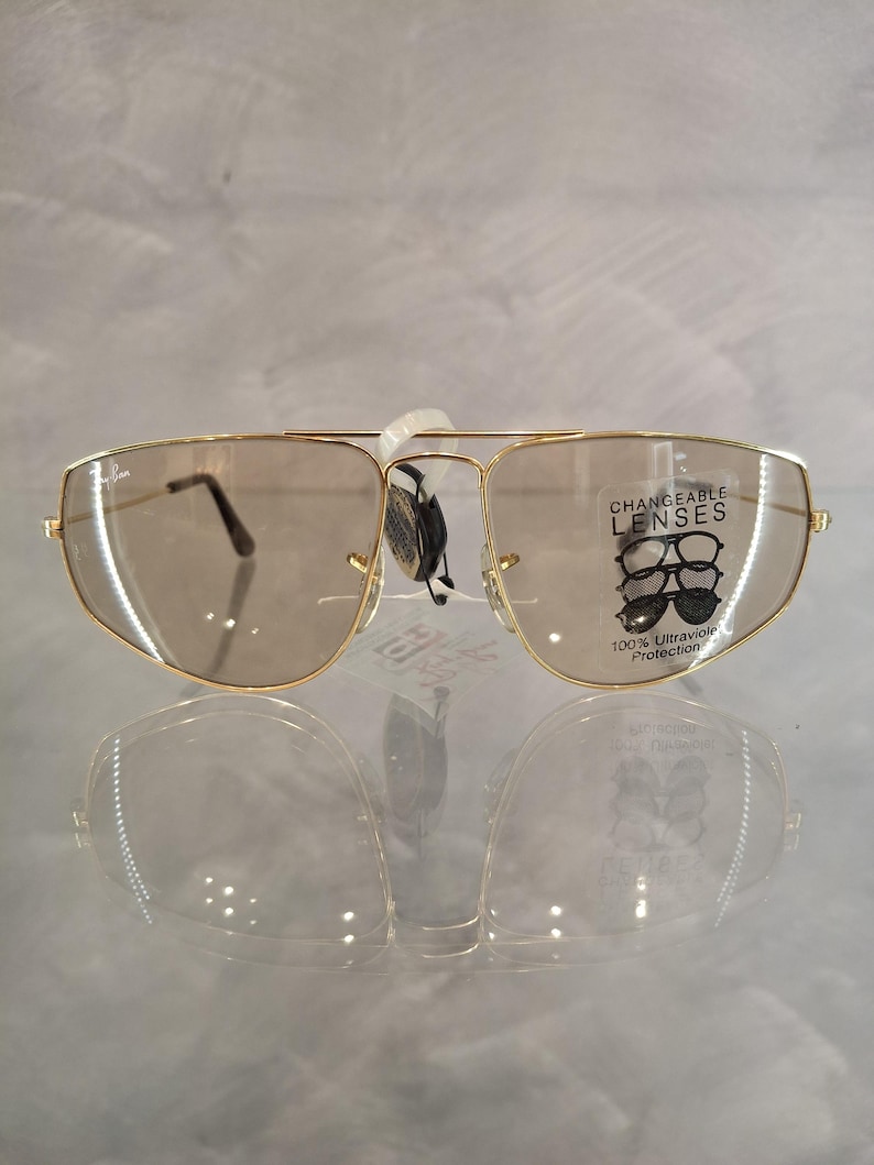May include: Gold-coloured sunglasses with brown lenses. The sunglasses have a double bridge and a rectangular shape. The lenses are marked with "CHANGEABLE LENSES" and "100% Ultraviolet Protection".