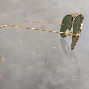 May include: Gold aviator sunglasses with green lenses. The sunglasses are folded and the arms are bent back.