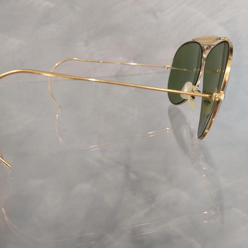 May include: Gold aviator sunglasses with green lenses. The sunglasses are folded and the arms are bent back.