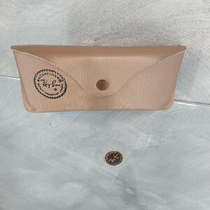 Puede incluir: Una funda de cuero marr&oacute;n para gafas de sol con el logotipo de Ray-Ban grabado en relieve en la parte delantera. La funda tiene un cierre de presi&oacute;n y un peque&ntilde;o bot&oacute;n de metal.