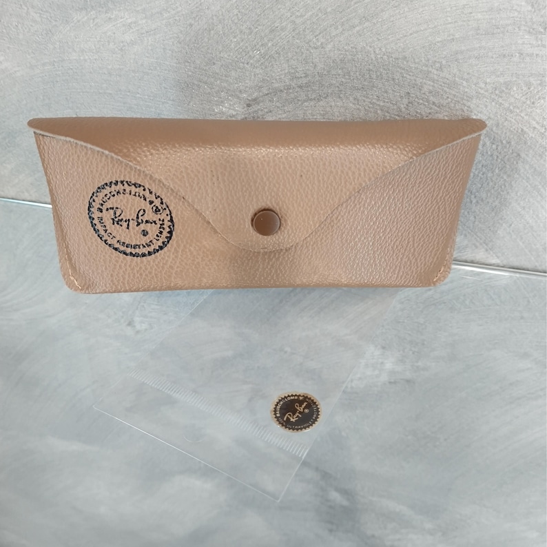 Puede incluir: Una funda de cuero marr&oacute;n para gafas de sol con el logotipo de Ray-Ban grabado en relieve en la parte delantera. La funda tiene un cierre de presi&oacute;n y un peque&ntilde;o bot&oacute;n de metal.