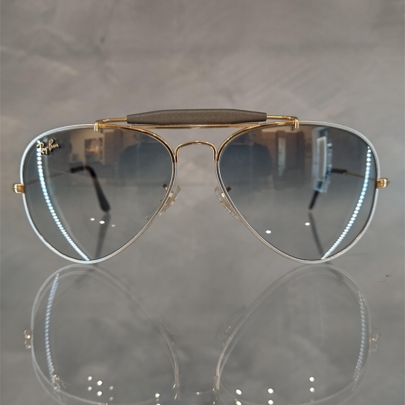 Puede incluir: Gafas de sol aviador doradas con puente gris y lentes azules degradados.