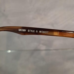 May include: Close-up of the arm of a pair of eyeglasses. The frame is a mottled brown color. The text "GATSBY STYLE 5 W0937" is printed in white on the arm of the glasses.