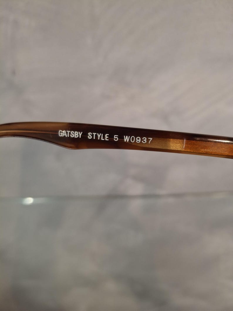 May include: Close-up of the arm of a pair of eyeglasses. The frame is a mottled brown color. The text "GATSBY STYLE 5 W0937" is printed in white on the arm of the glasses.
