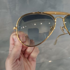 May include: Gold aviator sunglasses with blue tinted lenses. The sunglasses are being held in a hand.