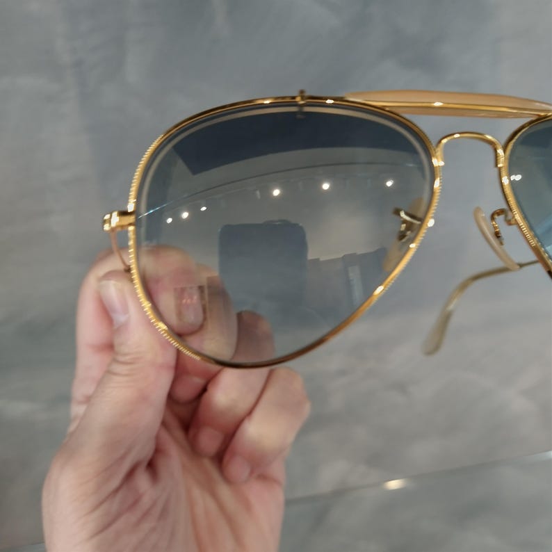 May include: Gold aviator sunglasses with blue tinted lenses. The sunglasses are being held in a hand.