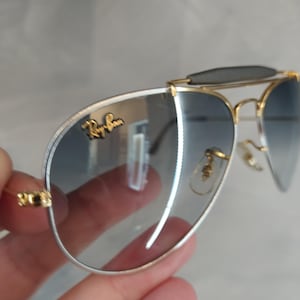 Puede incluir: Gafas de sol aviador con montura dorada y lentes azules degradados. Las gafas tienen una banda plateada alrededor de las lentes y el logotipo de Ray-Ban es visible en la lente derecha.