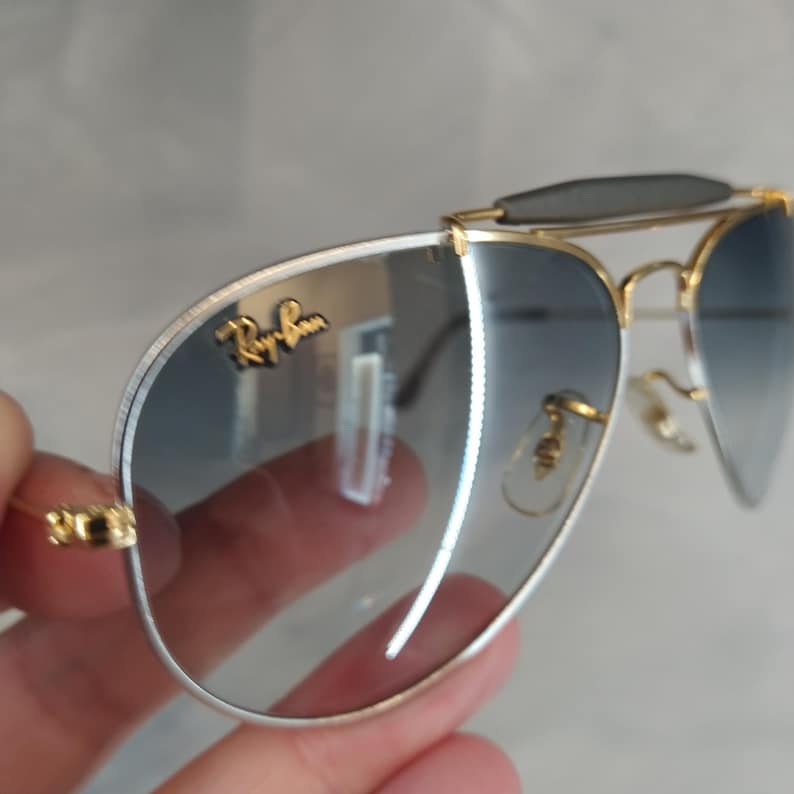 Puede incluir: Gafas de sol aviador con montura dorada y lentes azules degradados. Las gafas tienen una banda plateada alrededor de las lentes y el logotipo de Ray-Ban es visible en la lente derecha.