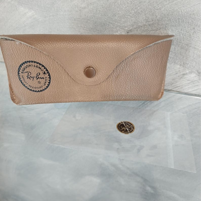 May include: A brown leather Ray-Ban sunglasses case with a gold metal snap closure. The case has a Ray-Ban logo with the text "Ray-Ban" and "Genuine Ray-Ban Sunglasses" in a circle.