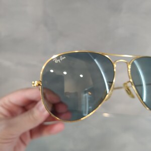 May include: Gold-framed aviator sunglasses with dark lenses. The Ray-Ban logo is visible on the upper left lens. The sunglasses have a classic teardrop shape and a thin gold metal frame.