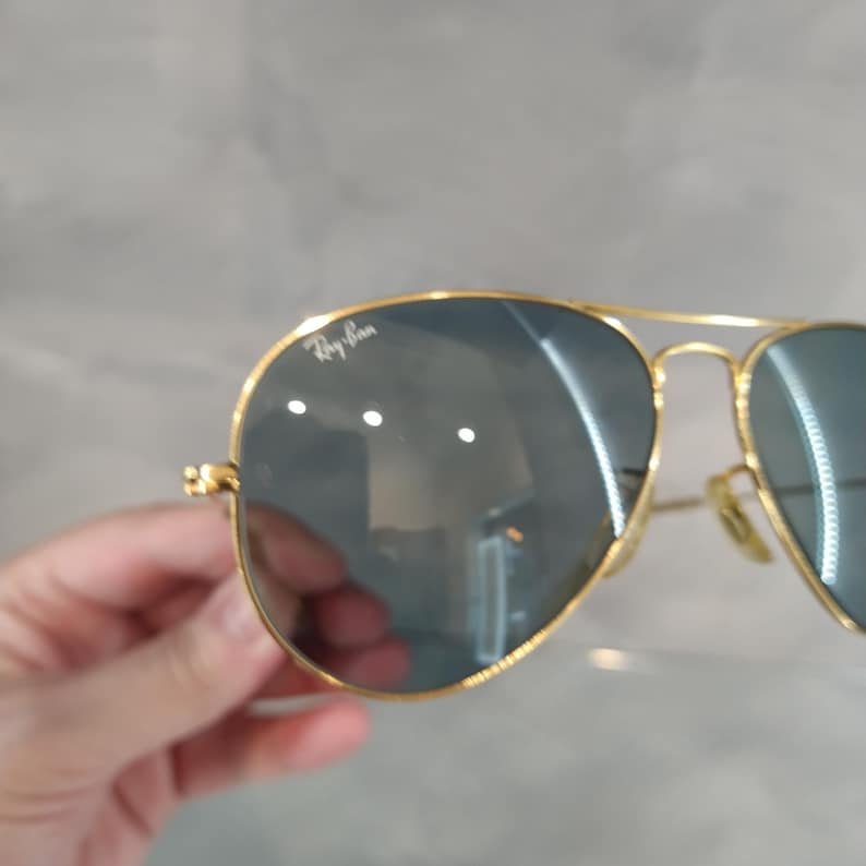 May include: Gold-framed aviator sunglasses with dark lenses. The Ray-Ban logo is visible on the upper left lens. The sunglasses have a classic teardrop shape and a thin gold metal frame.