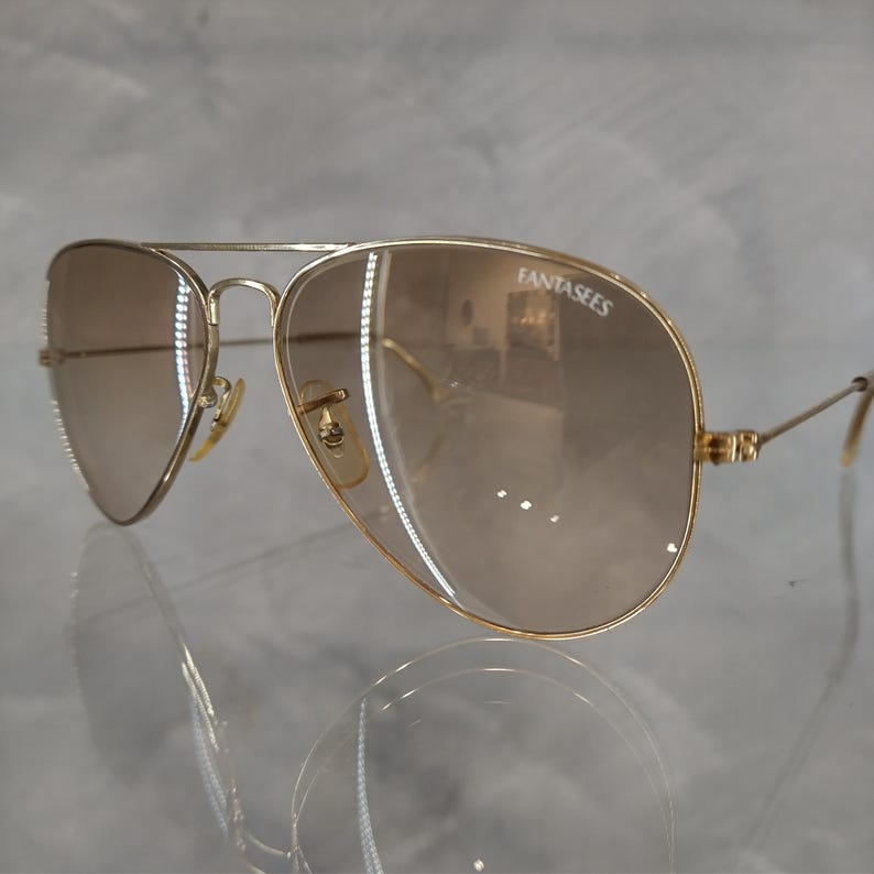 Puede incluir: Gafas de sol aviador doradas con lentes de degradado marr&oacute;n. Las gafas tienen la palabra "FantasEyes" impresa en la lente derecha.