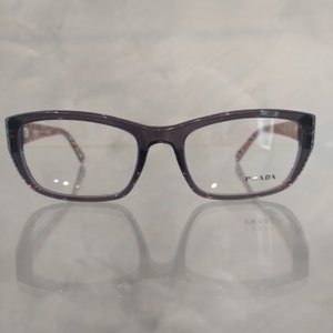 Könnte beinhalten: Graue Cat-Eye-Brille mit einem schwarz-weißen Muster auf der Innenseite der Bügel. Der Markenname "PRADA" ist auf dem rechten Bügel aufgedruckt.