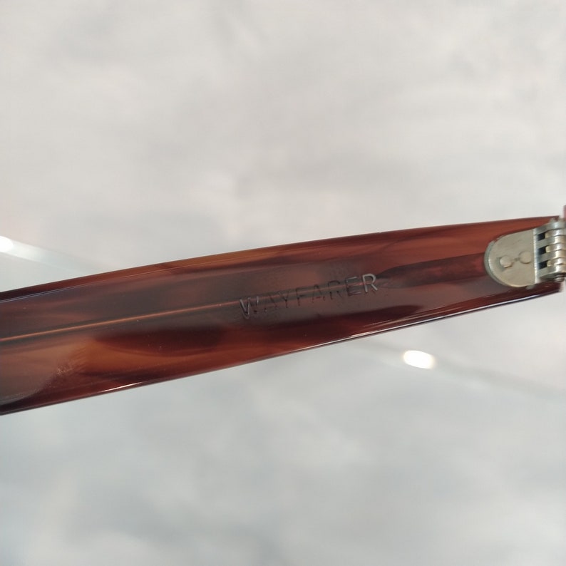 May include: Close-up of the brown plastic arm of a pair of Wayfarer sunglasses. The text "WAYFARER" is embossed on the arm.
