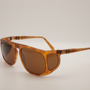 Może przedstawiać: Okulary przeciwsłoneczne w kolorze brązowego żółwia z brązowymi szkłami i osłoną boczną. Okulary mają metalowy detal zawiasu.