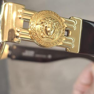 K&ouml;nnte beinhalten: Nahaufnahme einer goldfarbenen Sonnenbrille mit einem Medusenhaupt-Emblem am Scharnier. Die Sonnenbrille hat einen dunkelbraunen Rahmen und einen goldfarbenen Metallakzent. Das Medusenhaupt befindet sich in der Mitte eines kreisf&ouml;rmigen Designs.