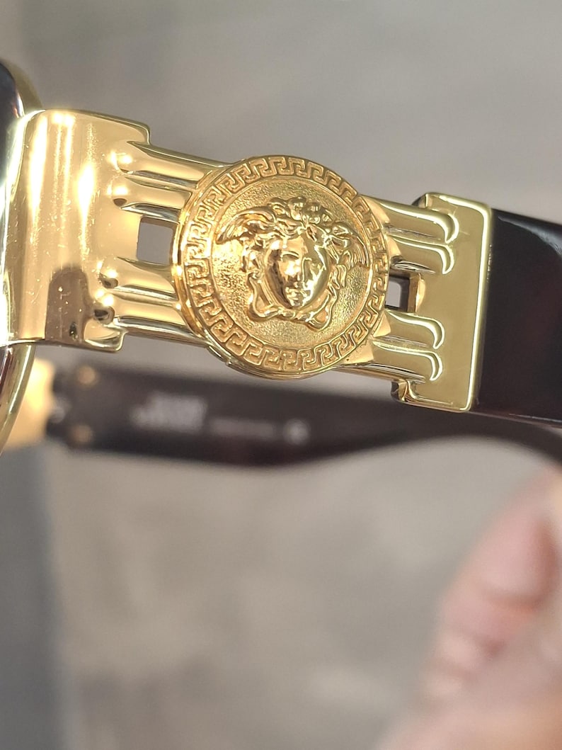 K&ouml;nnte beinhalten: Nahaufnahme einer goldfarbenen Sonnenbrille mit einem Medusenhaupt-Emblem am Scharnier. Die Sonnenbrille hat einen dunkelbraunen Rahmen und einen goldfarbenen Metallakzent. Das Medusenhaupt befindet sich in der Mitte eines kreisf&ouml;rmigen Designs.