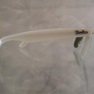 Pu&ograve; includere: Occhiali da sole bianchi con il logo Ray-Ban nero sull'asta. Gli occhiali hanno un design classico con montatura e aste spesse. Le lenti sono di colore scuro.