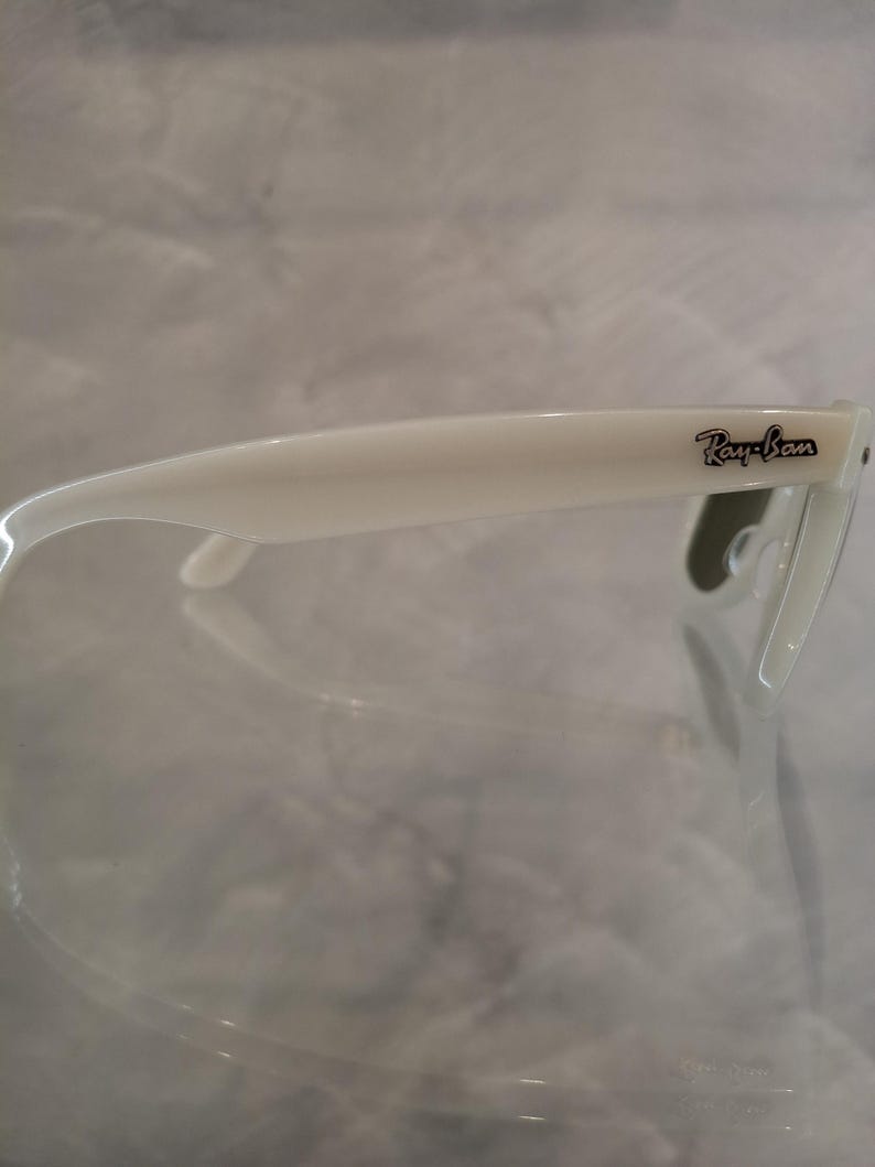 Pu&ograve; includere: Occhiali da sole bianchi con il logo Ray-Ban nero sull'asta. Gli occhiali hanno un design classico con montatura e aste spesse. Le lenti sono di colore scuro.