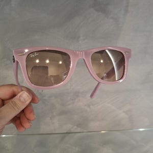 Puede incluir: Gafas de sol rosas con lentes marrones. El logotipo de Ray-Ban es visible en la esquina superior izquierda de la montura. Las gafas de sol est&aacute;n sujetas por una mano sobre un fondo gris.