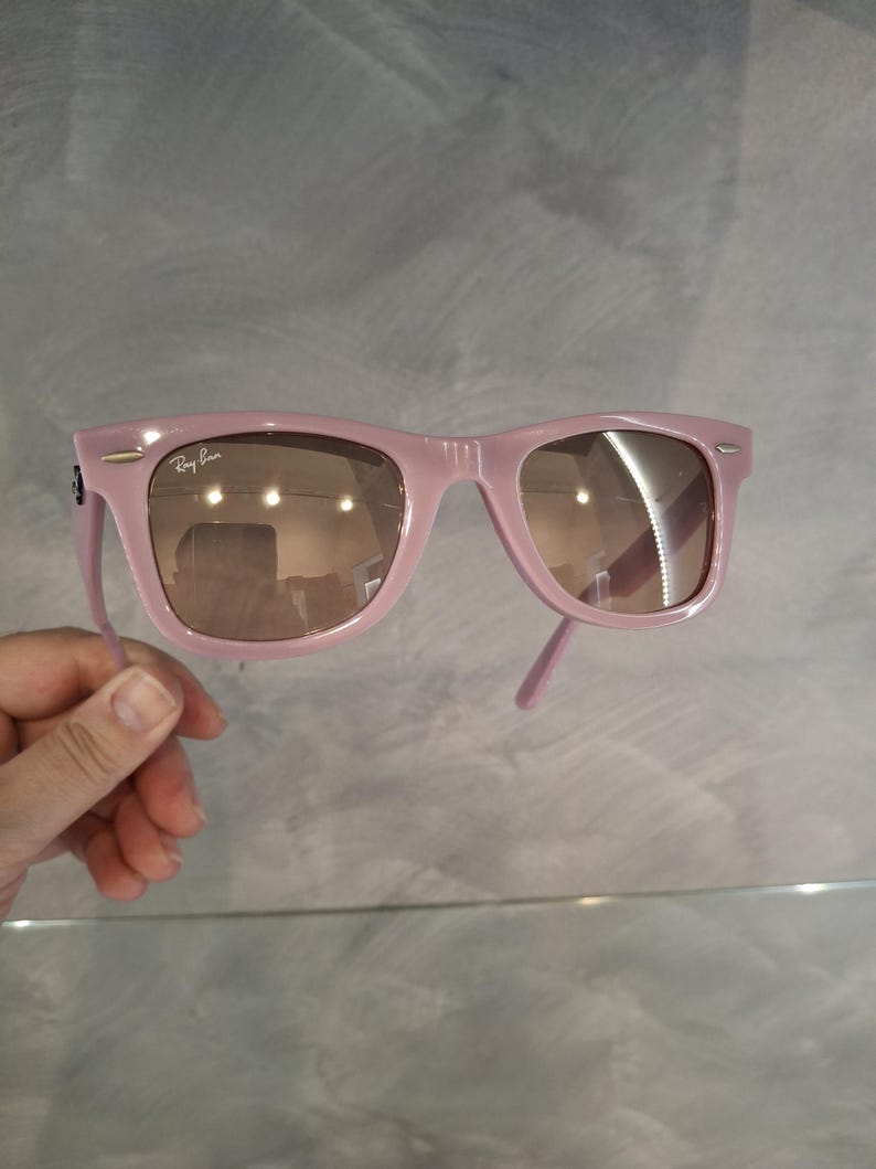 Puede incluir: Gafas de sol rosas con lentes marrones. El logotipo de Ray-Ban es visible en la esquina superior izquierda de la montura. Las gafas de sol est&aacute;n sujetas por una mano sobre un fondo gris.