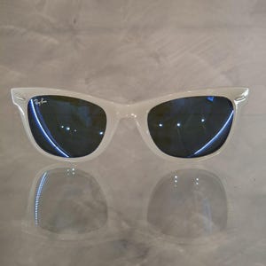 Pu&ograve; includere: Occhiali da sole bianchi con lenti blu scuro. Il logo Ray-Ban &egrave; visibile sulla lente sinistra. Gli occhiali da sole si riflettono sulla superficie sottostante.