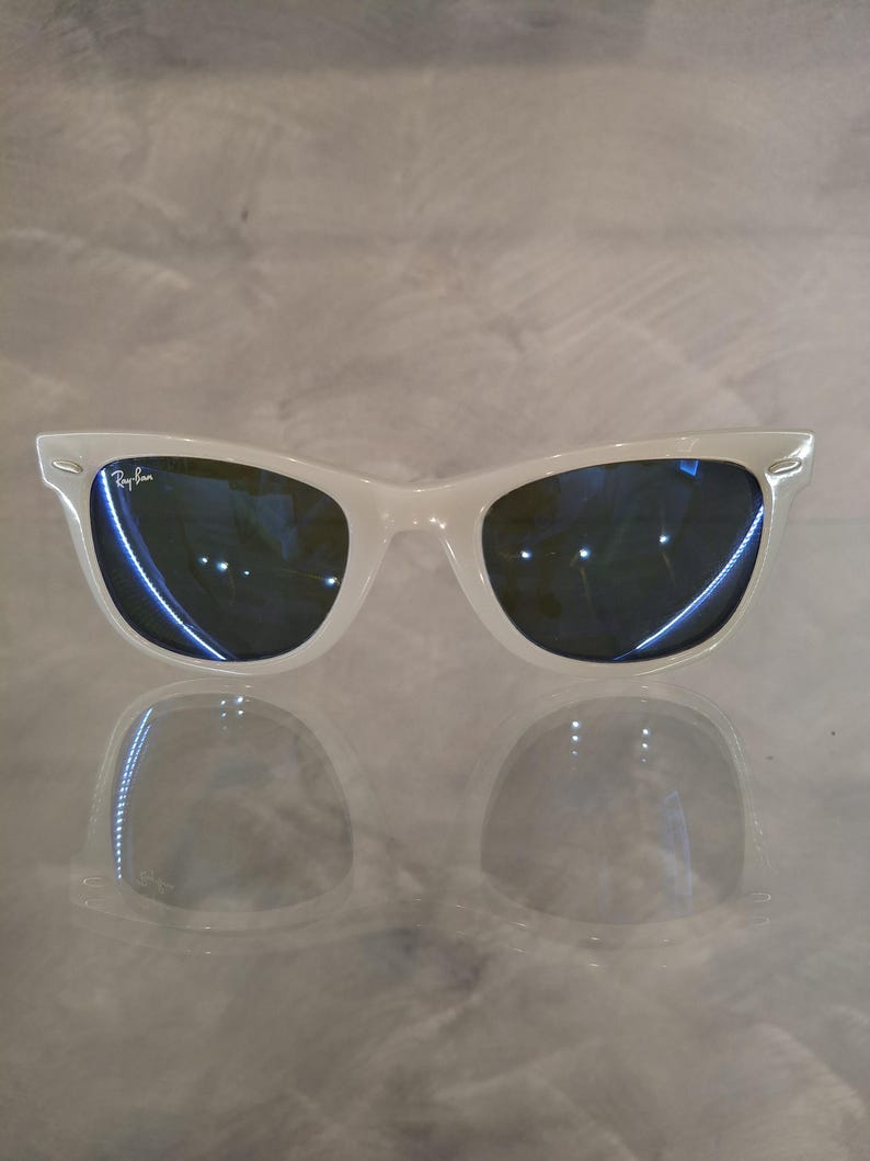 Pu&ograve; includere: Occhiali da sole bianchi con lenti blu scuro. Il logo Ray-Ban &egrave; visibile sulla lente sinistra. Gli occhiali da sole si riflettono sulla superficie sottostante.