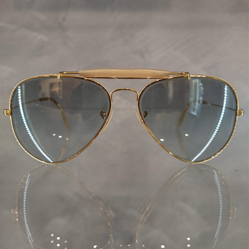 May include: Gold aviator sunglasses with a grey gradient lens. The sunglasses have a thin gold metal frame and a brown bridge.