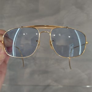 Puede incluir: Gafas de sol cuadradas con montura dorada y lentes tintados de azul.
