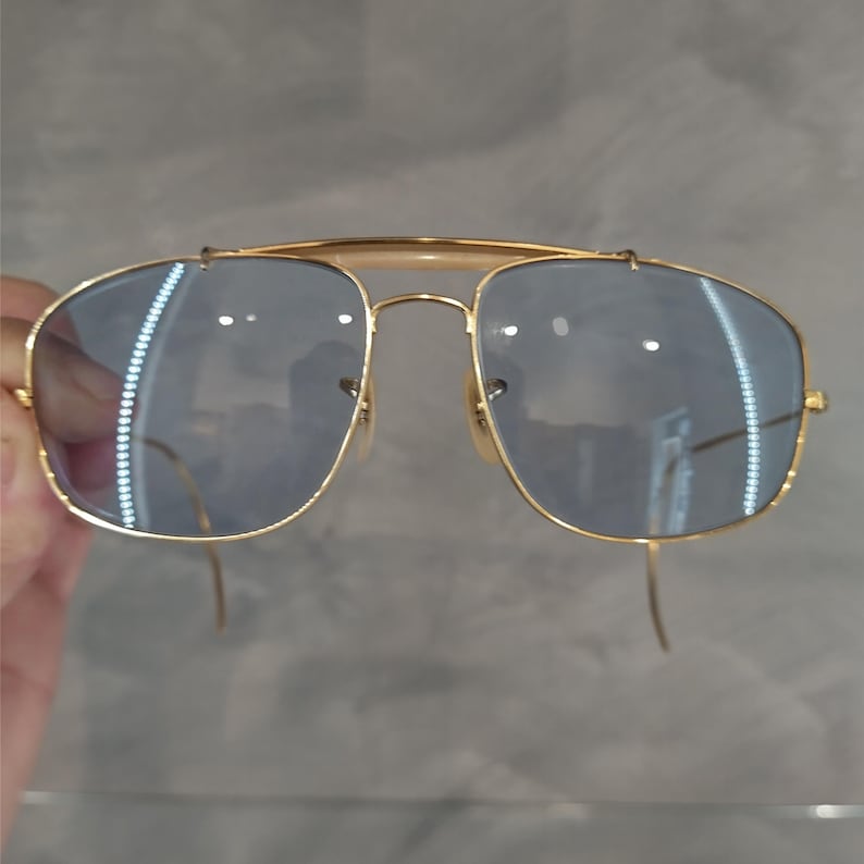 Puede incluir: Gafas de sol cuadradas con montura dorada y lentes tintados de azul.
