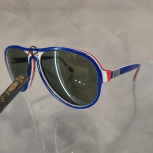 May include: Blue, white, and red aviator sunglasses with a Ray-Ban logo on the side. The sunglasses have a green lens.