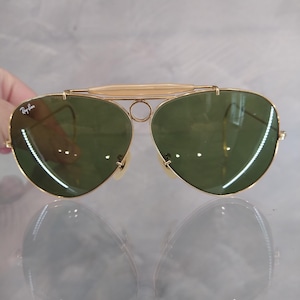 May include: Gold aviator sunglasses with green lenses and a Ray-Ban logo on the left lens.
