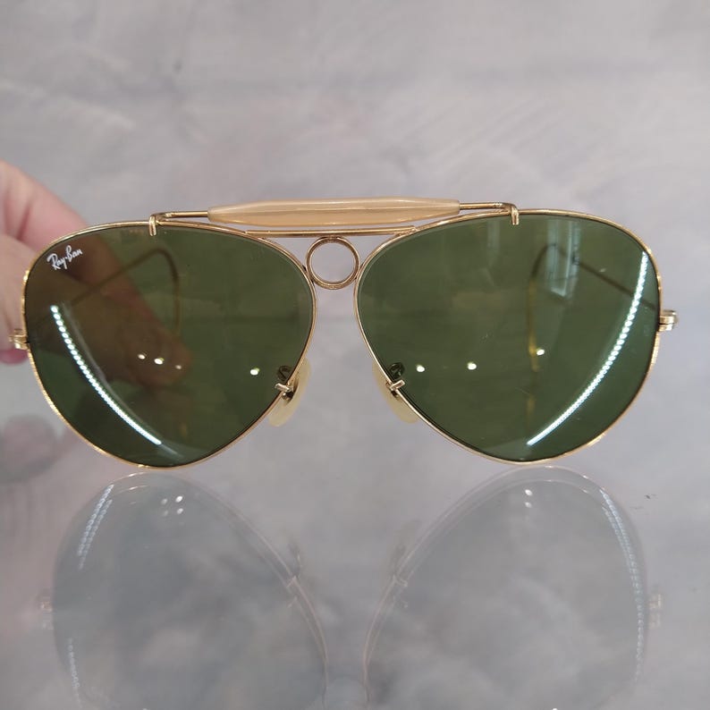 May include: Gold aviator sunglasses with green lenses and a Ray-Ban logo on the left lens.