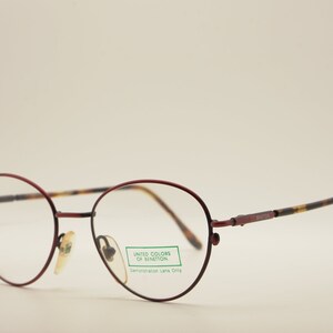 Puede incluir: Gafas de metal con montura roja y lentes redondas. La montura tiene un patrón de carey en las patillas. La etiqueta en la montura dice "United Colors of Benetton" y "Demonstration Lens Only".