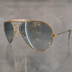 May include: Gold aviator sunglasses with a white bar across the bridge and blue gradient lenses.