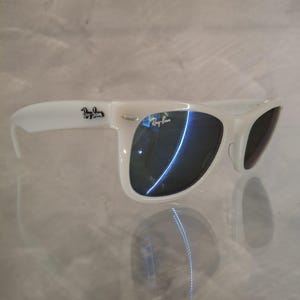 Pu&ograve; includere: Occhiali da sole bianchi con lenti scure. Il logo Ray-Ban &egrave; visibile sulla montatura. Gli occhiali da sole sono appoggiati su una superficie riflettente.