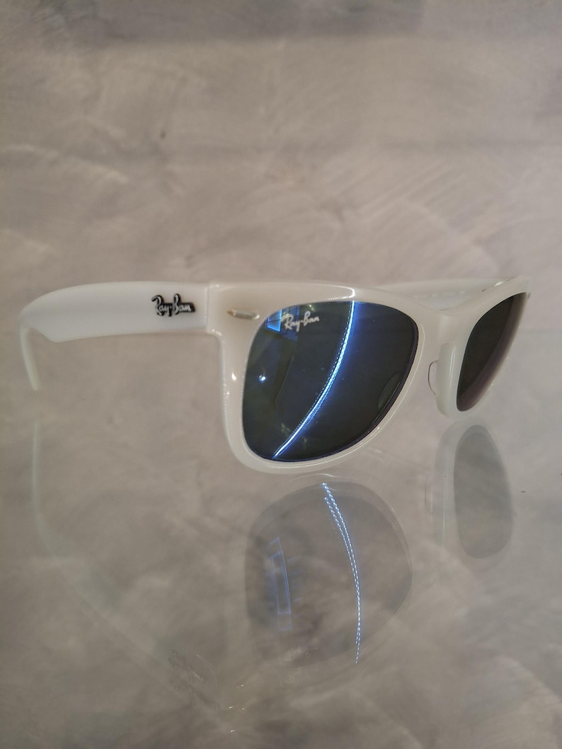 Pu&ograve; includere: Occhiali da sole bianchi con lenti scure. Il logo Ray-Ban &egrave; visibile sulla montatura. Gli occhiali da sole sono appoggiati su una superficie riflettente.