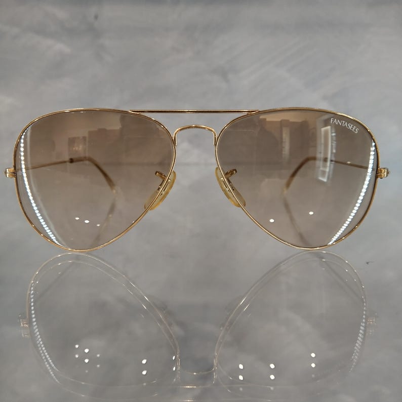 Puede incluir: Gafas de sol aviador doradas con lentes de degradado marr&oacute;n. Las gafas tienen la palabra "Fantasies" impresa en la lente derecha.
