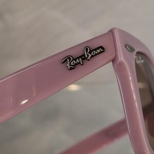 Puede incluir: Primer plano de unas gafas de sol Ray-Ban rosas. Las monturas son de color rosa liso y brillante con el logotipo de Ray-Ban plateado en la patilla. Las lentes son de color marr&oacute;n claro.