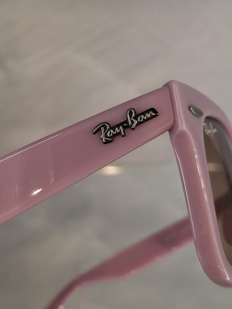 Puede incluir: Primer plano de unas gafas de sol Ray-Ban rosas. Las monturas son de color rosa liso y brillante con el logotipo de Ray-Ban plateado en la patilla. Las lentes son de color marr&oacute;n claro.