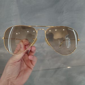 Puede incluir: Gafas de sol aviador doradas con lentes de degradado marr&oacute;n. Las gafas tienen la palabra "Fantasie" impresa en la lente derecha.