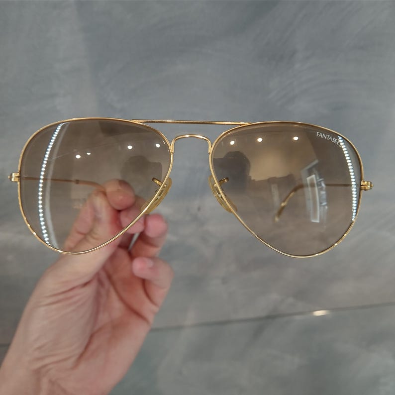 Puede incluir: Gafas de sol aviador doradas con lentes de degradado marr&oacute;n. Las gafas tienen la palabra "Fantasie" impresa en la lente derecha.